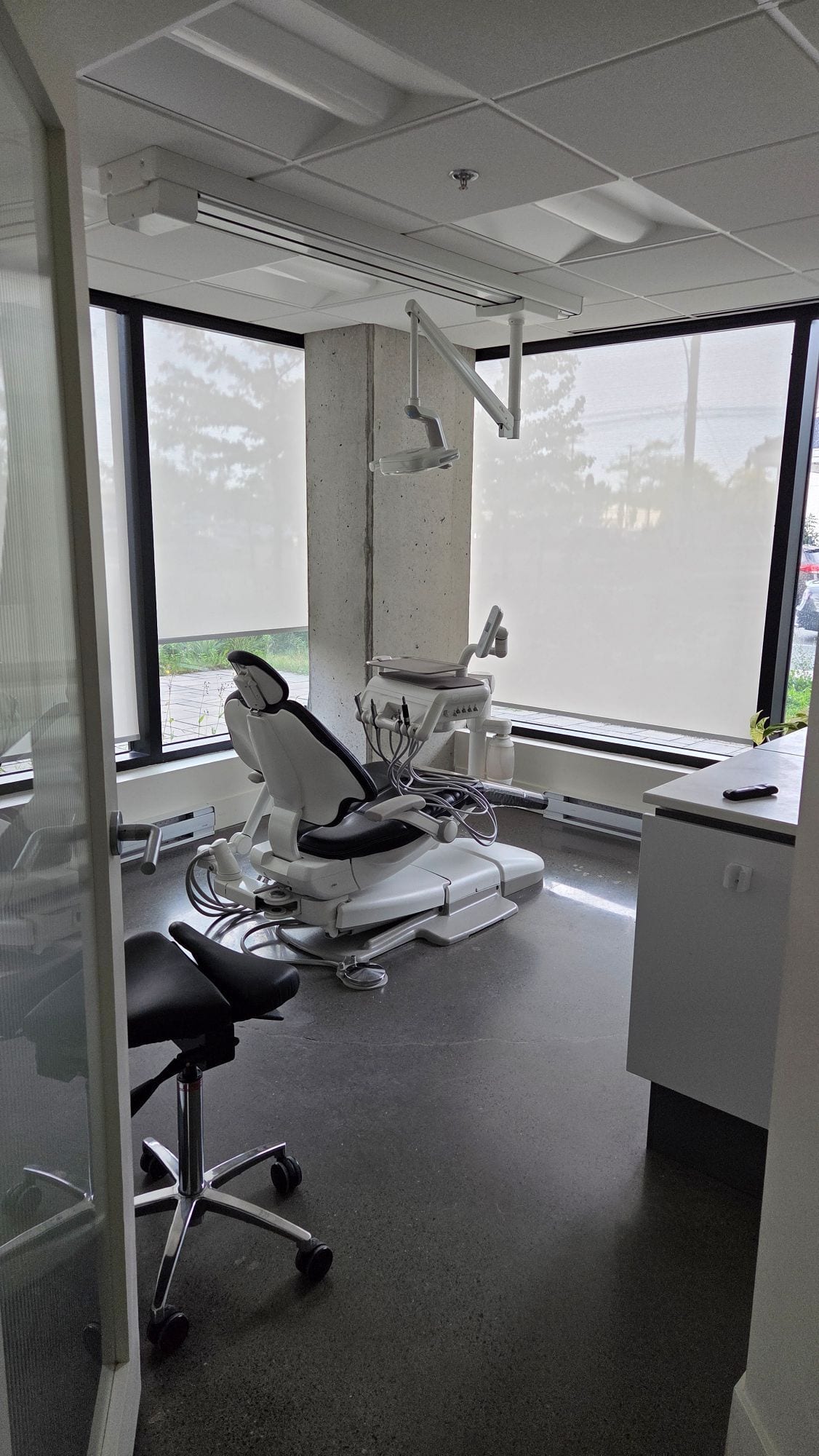 Salle de traitement vue depuis la porte : stores enroulables baissés devant de grandes fenêtres d'angle, fauteuil dentaire vide
