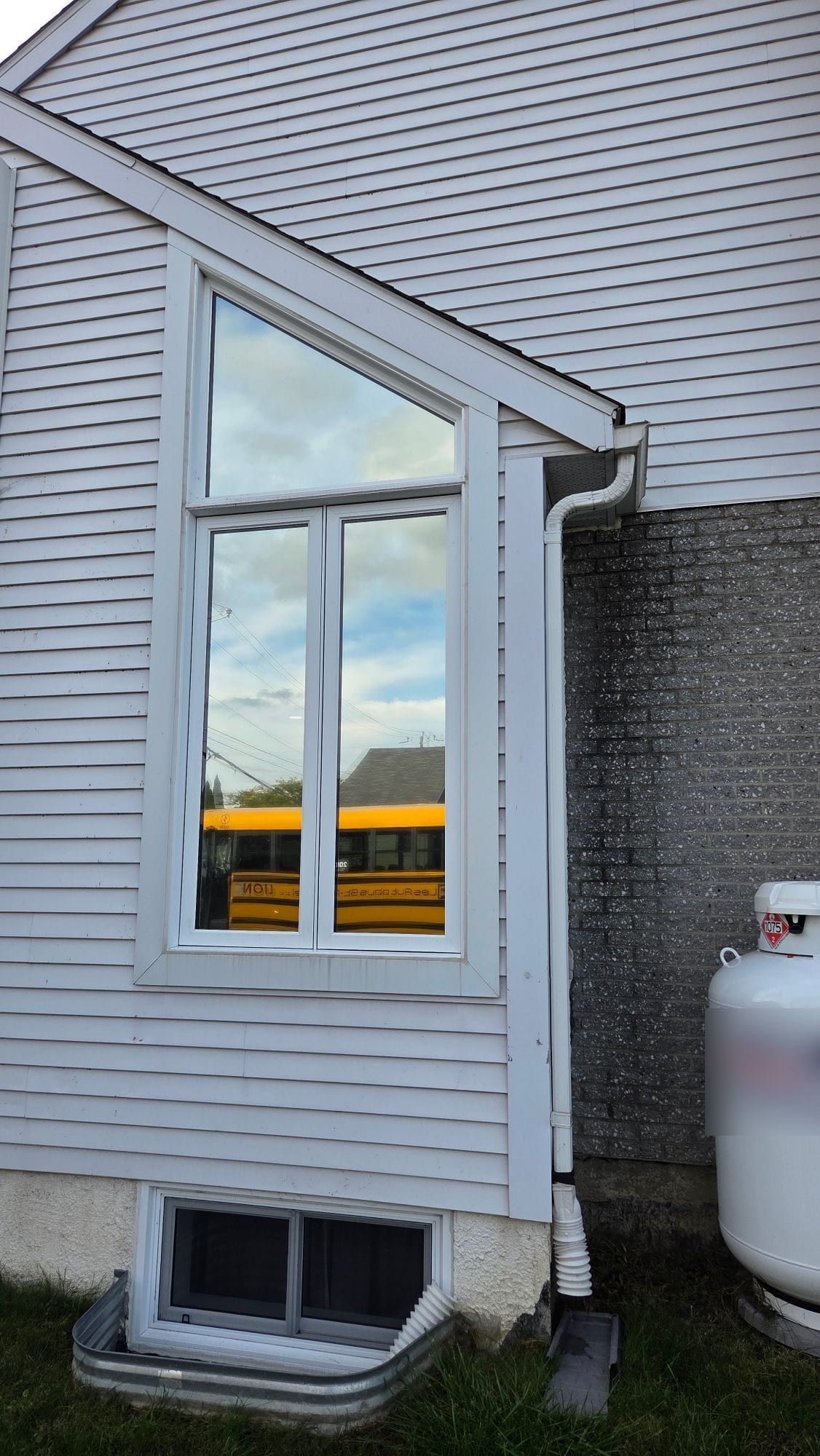 La fenêtre à sommet triangulaire vue de l’extérieur : la vitre renvoie le ciel et un autobus scolaire